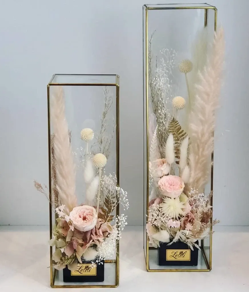 The image showcases two elegant, rectangular glass vases filled with a delicate arrangement of dried and preserved flowers by Lelili Fleurs, a wedding florist in the Mariage Elegant vendor listing, Canada. Each vase contains carefully styled pampas grass, soft pink roses, white globe amaranth, and baby's breath, creating an exquisite, romantic look suitable for wedding decor. The floral arrangements are set against a crisp white background, emphasizing their intricate beauty and timeless charm.