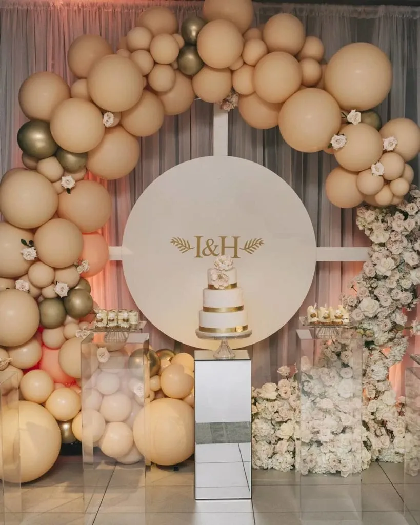 The image showcases a beautifully arranged wedding setup by Lelili Fleurs, categorized under Wedding Florists. It features an elegant circular backdrop with the initials 'I & H' centered, surrounded by an arch of cream and gold balloons. Below, a multi-tiered white wedding cake with gold accents is displayed on a sleek mirrored pedestal. Flanking the cake are glass stands adorned with delicate white flowers. Abundant floral arrangements of white roses and greenery decorate the base, enhancing the sophisticated and romantic atmosphere.