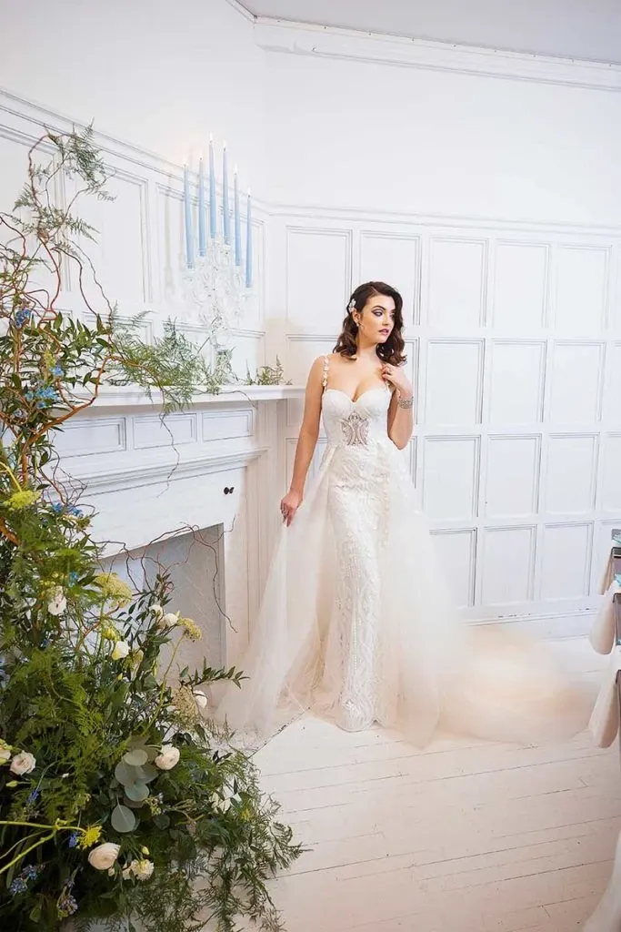 In this image, a bride stands elegantly in a luxurious white room, showcasing a stunning lace wedding gown with intricate detailing. She is positioned near a white mantel adorned with light blue candles, adding a soft, romantic ambiance. To her left, a lush floral arrangement by Lelili Fleurs, under the wedding florists category, features a mix of greenery and delicate white flowers, perfectly complementing the bridal theme. The setting exudes an atmosphere of sophistication and grace, ideal for a tasteful wedding celebration.
