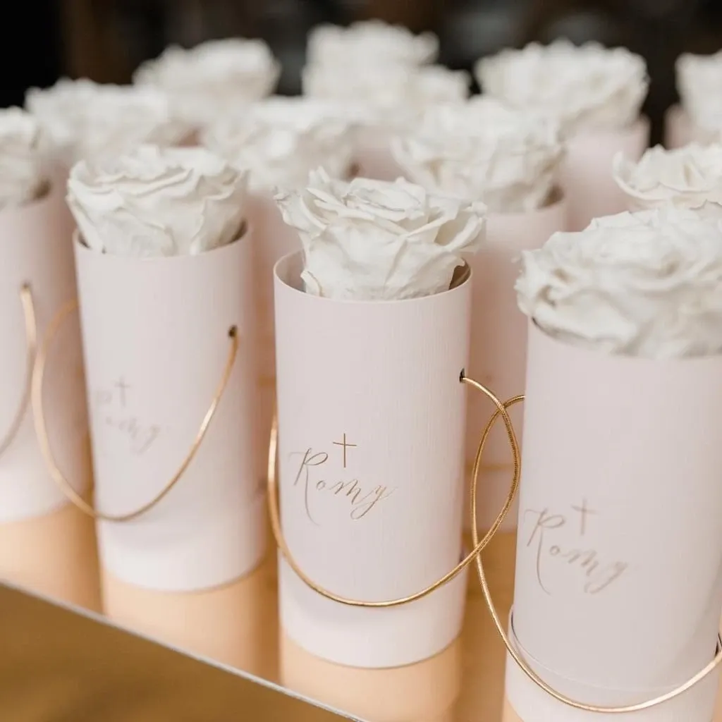 The image showcases elegant floral arrangements by Lelili Fleurs, a distinguished wedding florist, for Mariage Elegant in Canada. Each arrangement consists of a single white rose beautifully presented in a blush pink cylindrical box, embellished with the word 'Romy' and a delicate cross in gold. These floral favors are neatly arranged in rows, highlighting their refined and luxurious design, perfect for a sophisticated wedding ambiance.