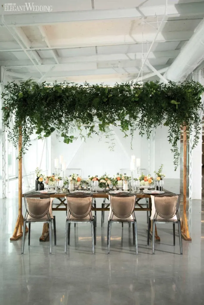 In this image, a beautifully arranged wedding table by Lelili Fleurs is showcased under an elegant canopy of lush greenery and cascading vines. The table setting, designed by the wedding florists, features a centerpiece adorned with an assortment of pastel flowers, including roses and peonies, placed amongst candles in glass holders. The decor exudes a sophisticated ambiance, enhanced by the soft glow of overhead lighting. Six modern, upholstered chairs are neatly lined up along the table, reflecting the elegant wedding theme. This setup, captured in a spacious venue with high ceilings, embodies the exquisite floral artistry of Lelili Fleurs, a renowned wedding florist in Canada, perfect for creating unforgettable wedding atmospheres.