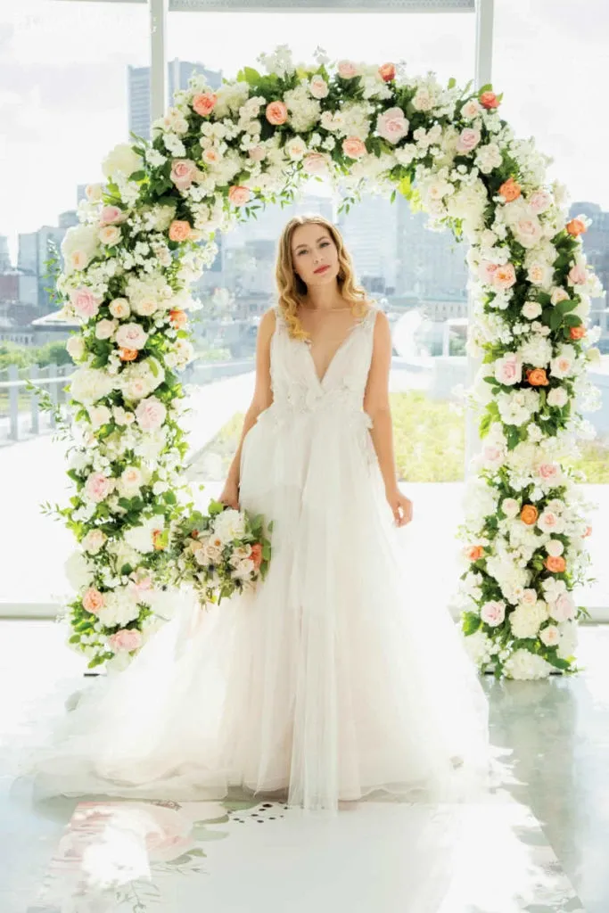 A bride stands in front of a stunning floral arch designed by Lelili Fleurs, a renowned vendor in the Wedding Florists category. The arch is beautifully adorned with white, pink, and peach flowers, creating an elegant and romantic backdrop. The bride is wearing a flowing white gown with intricate lace details, holding a bouquet that complements the floral arch. The background features a bright, sunlit cityscape, adding a modern touch to the classic wedding scene.