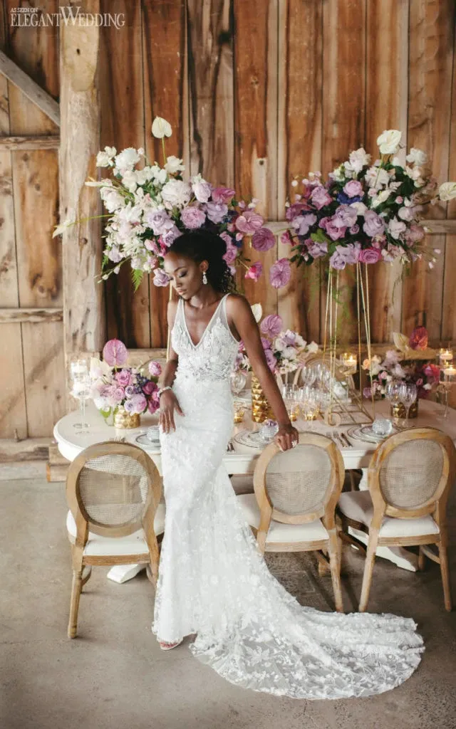 A bride in a white lace wedding gown stands gracefully beside an elegantly decorated table. The table is adorned with luxurious floral arrangements by Lelili Fleurs, featuring a mix of pink, purple, and white flowers. These vibrant blossoms, set against a rustic wooden backdrop, showcase the vendor's expertise as a wedding florist. The soft candlelight and classic wooden chairs add a touch of romance to the setting, highlighting the exquisite details suitable for a refined, elegant wedding.