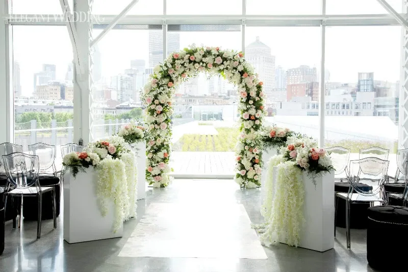 The image showcases a beautifully arranged wedding setup by Lelili Fleurs, a vendor in the Wedding Florists category. The scene is set in a bright, modern space with large windows offering a view of a city skyline. A stunning floral arch, adorned with pink and white roses mixed with lush greenery, is the centerpiece, creating a romantic and elegant backdrop for the ceremony. Two white pedestals are placed on either side, each cascading with floral arrangements that complement the arch. The arrangement reflects a blend of classic and contemporary design, ideal for sophisticated weddings.