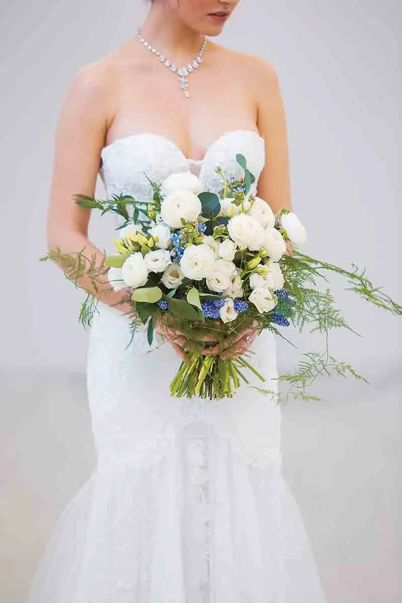 A bride in a strapless white lace wedding dress holds an elegant floral bouquet designed by Lelili Fleurs, categorized under Wedding Florists. The bouquet features a harmonious blend of white roses, lush greenery, and delicate blue accents, creating a classic yet fresh arrangement. The bride also wears a sparkling necklace, complementing the sophisticated and romantic aesthetic.