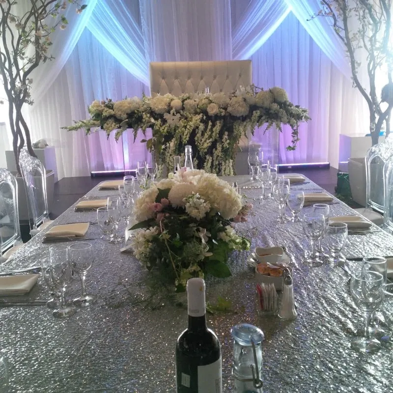 The image features an elegant wedding floral arrangement by La Vie Fleurie, a renowned wedding florist, at Mariage Elegant in Canada. The table is beautifully adorned with a shimmering silver tablecloth, surrounded by spotless glassware and neatly folded napkins. A stunning centerpiece composed of white and blush roses, lush greenery, and cascading floral elements is prominently displayed. In the background, a plush, tufted backdrop is elegantly framed by soft, draped curtains and tasteful branches, creating a sophisticated and romantic ambiance suitable for a wedding reception.