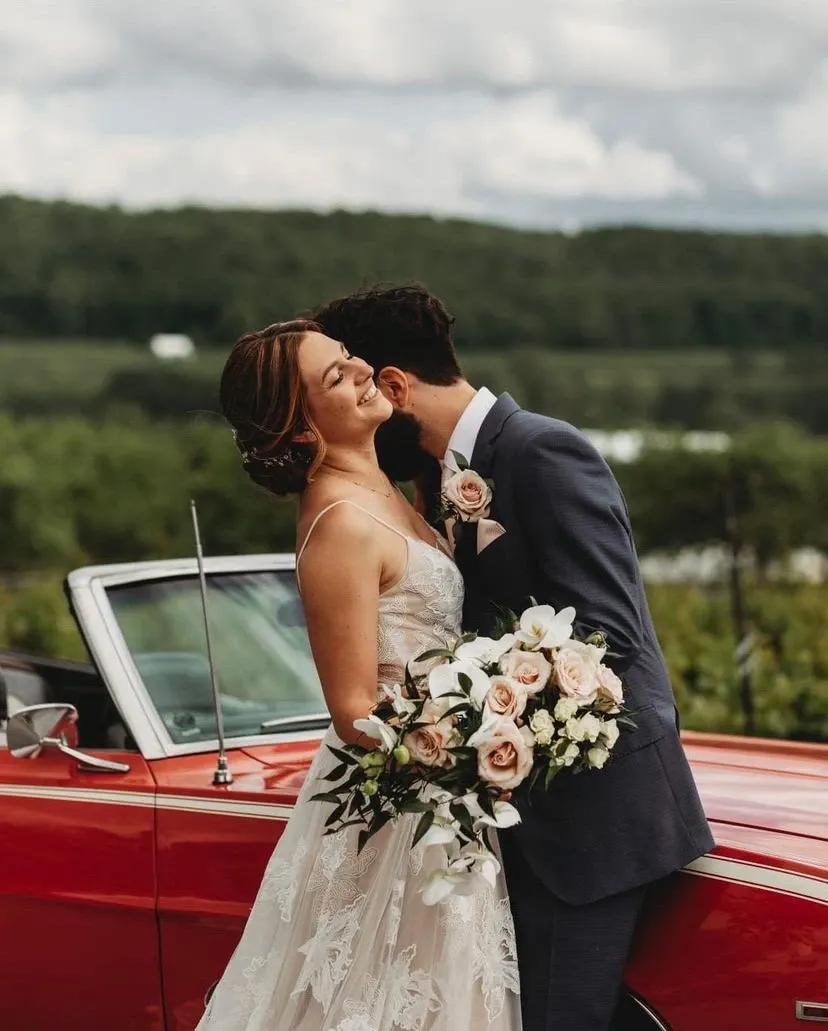 A joyful bride holding a bouquet of soft pink and white roses, expertly arranged by La Vie Fleurie, a wedding florist featured on Mariage Elegant, Canada. The couple stands beside a vintage red convertible against a lush, green landscape, evoking a romantic and elegant outdoor wedding atmosphere. The groom, in a stylish suit, leans in affectionately as they celebrate their special day.