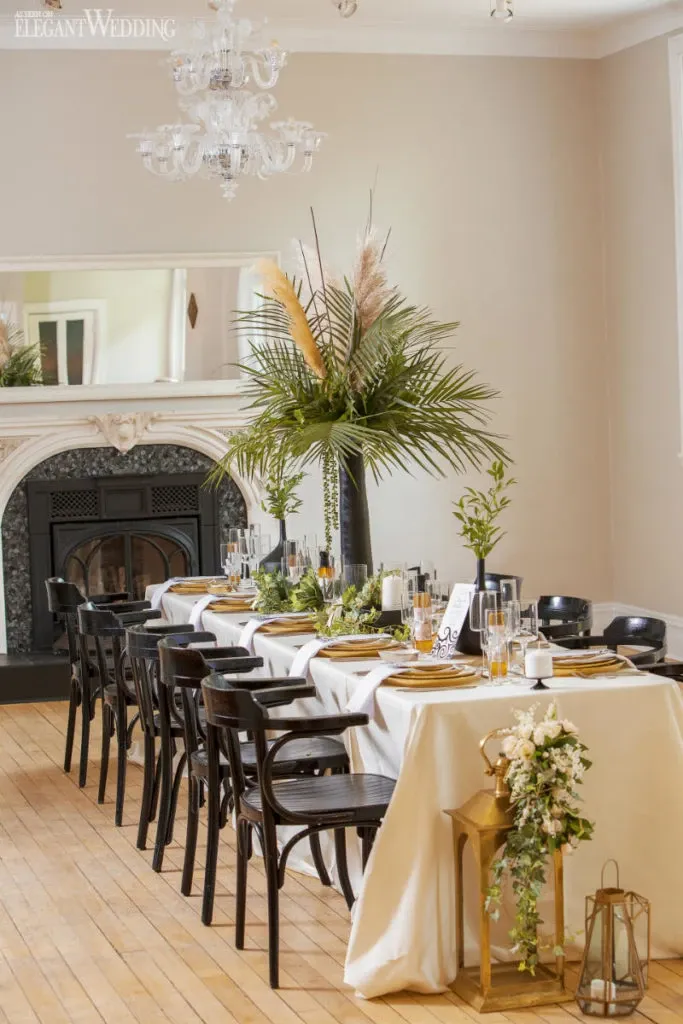 This image showcases an elegantly decorated wedding reception table by La Vie Fleurie, categorized under Wedding Florists. The table is adorned with lush green floral arrangements, featuring tall palm fronds and pampas grass at the centerpiece. The setting includes black chairs and a white tablecloth, complemented by gold accents such as cutlery and decorative lanterns. A grand chandelier hangs above, and the room boasts a sophisticated ambiance with a large mirror over the ornate fireplace, enhancing the elegant atmosphere suitable for a wedding event.
