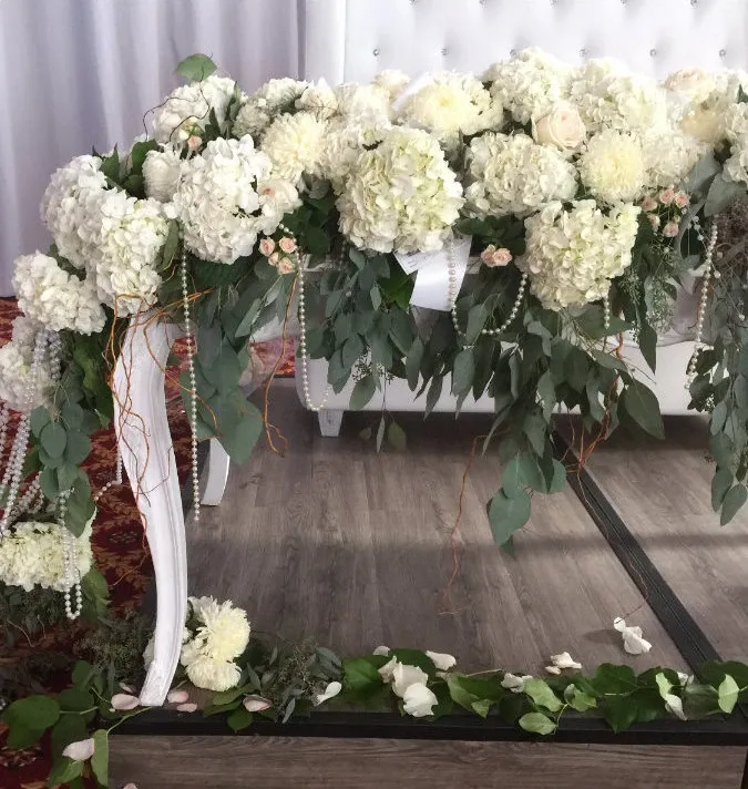 This image showcases an elegant floral arrangement by La Vie Fleurie, a wedding florist listed on Mariage Elegant, Canada. The arrangement features a cascading display of lush white hydrangeas and chrysanthemums, interspersed with soft pink roses. Delicate greenery and trailing vines add depth and texture, draping beautifully over the edges of a classic white table. Pearl strands are tastefully integrated, enhancing the romantic and sophisticated atmosphere. This exquisite setup reflects the floral artistry and attention to detail that La Vie Fleurie provides for weddings. The floral arrangement is set against a neutral backdrop and rests on a polished wooden floor, completing the serene and elegant wedding decor.