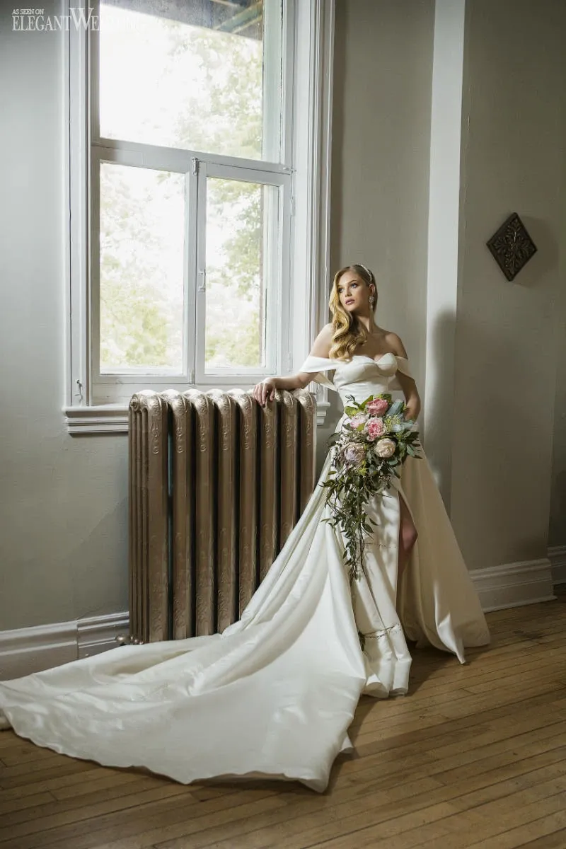 A bride stands elegantly in a softly lit room beside a vintage radiator, holding a cascading bouquet designed by La Vie Fleurie, a premier wedding florist listed on Mariage Elegant, Canada. The bouquet features lush pink and white flowers combined with trailing greenery, perfectly complementing the bride's off-the-shoulder, floor-length white gown with a long train. Natural light streams in through a large window, casting gentle shadows and highlighting the delicate details of the floral arrangement and bridal attire.