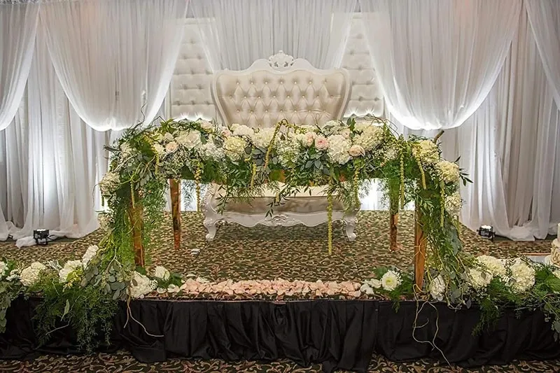 The image showcases an elegant wedding floral arrangement by La Vie Fleurie, a renowned wedding florist. The setting features a luxurious backdrop of white draped curtains and a sophisticated tufted white chair, exuding a sense of romance and grace. In the foreground, there's a beautifully decorated floral arch with cascading greenery and an assortment of white and pale pink flowers, creating a stunning focal point for a wedding ceremony or reception. The floral design reflects a classic and timeless aesthetic, perfect for a sophisticated and elegant wedding event in Canada.