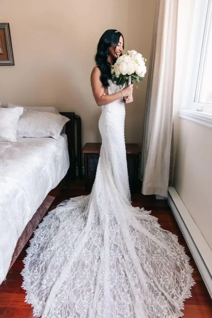 A bride, wearing an elegant lace wedding gown with a long, intricate train, stands in a softly lit room beside a bed. She is holding a beautifully arranged bouquet of white flowers crafted by La Vie Fleurie, showcasing their expertise as a wedding florist. The bride's dark hair cascades in soft waves, and she gazes contently out of a large window, enhancing the serene and romantic atmosphere. This image highlights the exquisite floral design perfect for weddings, offered by La Vie Fleurie, a featured vendor on Mariage Elegant, Canada.