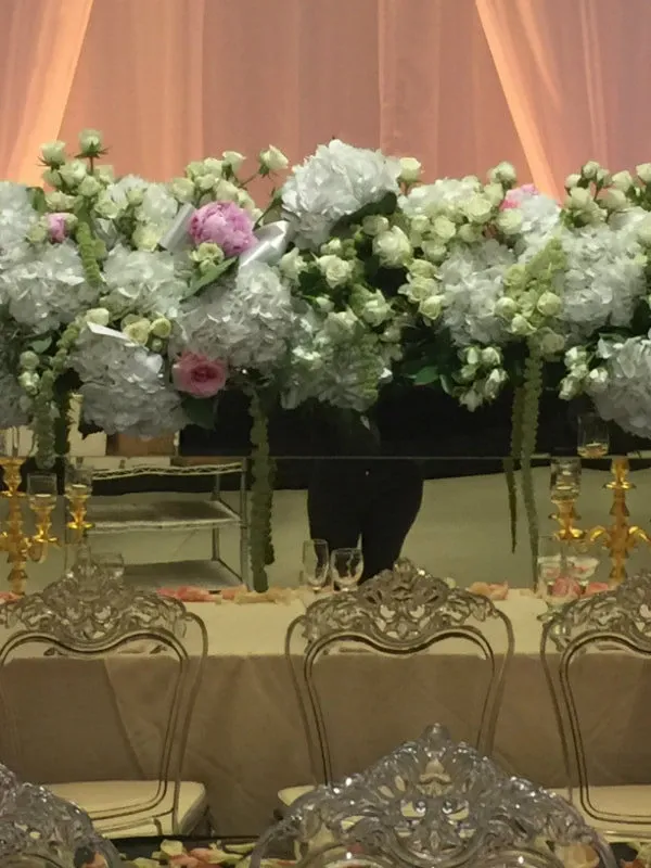 The image features an elegant floral arrangement by La Vie Fleurie, a wedding florist, displayed on a table setting suitable for a wedding reception. The arrangement includes an abundance of white and pink flowers, including hydrangeas and roses, artistically cascading with greenery. The display is elevated on golden stands, adding a touch of sophistication. In the background, sheer pink drapes create a soft and romantic ambiance. The table is adorned with ornate, transparent chairs, complementing the luxurious theme, and scattered rose petals add the final touch to this exquisite wedding setting by La Vie Fleurie.