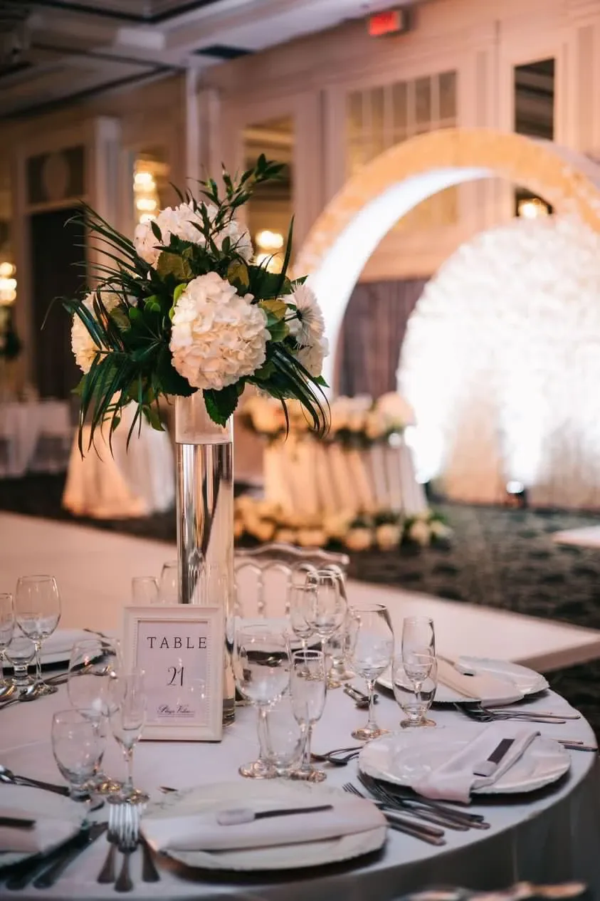 This image showcases an elegant wedding reception setup by La Vie Fleurie, a wedding florist under the category of Wedding Florists. A beautifully arranged floral centerpiece featuring white hydrangeas and lush greenery is placed in a tall, clear vase on a round table. The table is set with white tablecloth, neatly arranged cutlery, and glassware, along with a numbered table card marked 'Table 21.' In the background, a beautifully lit, circular floral arch adds to the romantic ambiance, making it a perfect setting for an elegant wedding celebration in Canada.