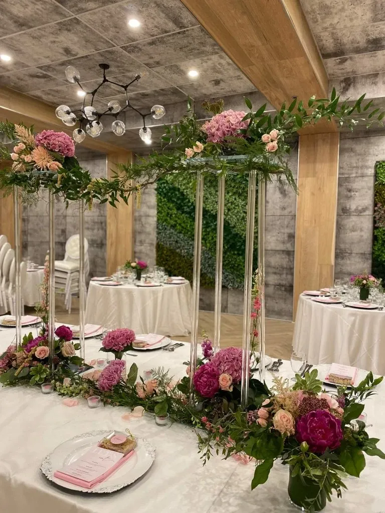 This image features an elegant wedding reception setup designed by La Vie Fleurie, a renowned wedding florist in the category of wedding florists on Mariage Elegant, Canada. The arrangement includes lush floral centerpieces with vibrant pink and deep magenta flowers atop tall, slender glass stands, accented with sprigs of fresh greenery. The tables are adorned with white tablecloths, fine china, and pink menus, creating a cohesive and sophisticated look. The background showcases a modern venue with rustic wooden beams and a green wall, enhancing the natural and elegant atmosphere of the wedding setting.