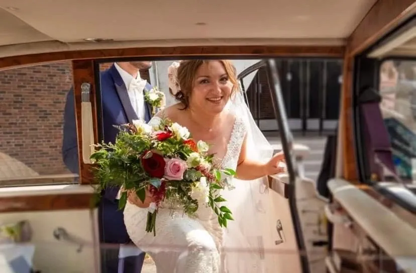 A bride joyfully steps into a classic car while holding a stunning bouquet crafted by La Vie Fleurie, a renowned wedding florist featured on Mariage Elegant, Canada. The bouquet includes a mix of red, pink, and white flowers with lush green foliage, reflecting the artistry and elegance of La Vie Fleurie's floral designs. In the background, the groom, dressed in a sharp blue suit with a white boutonniere, is partially visible, enhancing the celebratory moment. The image captures the excitement and elegance of a well-planned wedding celebration.