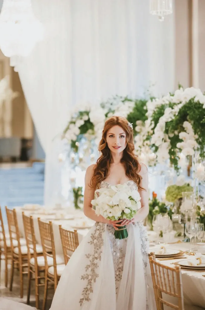In this image, a bride stands elegantly in a beautifully decorated wedding venue by Alain Simon Fleurs, highlighting their expertise as a wedding florist. She is holding a lush bouquet of white roses and greenery, which complements her detailed, flowing white gown. The background features a long table adorned with an exquisite floral arrangement of white flowers and greenery. The table is set with fine dining ware, and gold chiavari chairs add a touch of sophistication to the setting, exemplifying the high-quality floral services provided by Alain Simon Fleurs for weddings in Canada.