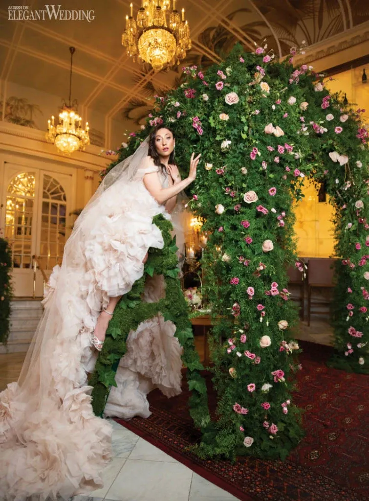 This image features a beautifully arranged floral arch crafted by Alain Simon Fleurs, showcasing their expertise as a wedding florist. The arch is adorned with lush greenery and a variety of roses in soft pink and cream hues, creating a romantic and elegant setting. A bride in a voluminous, ruffled wedding gown is gracefully posing next to the arch, enhancing the luxurious wedding ambiance. The background includes opulent chandeliers and classic architectural details, emphasizing the sophistication of the venue. This image highlights the exceptional floral design capabilities of Alain Simon Fleurs in creating memorable wedding settings.