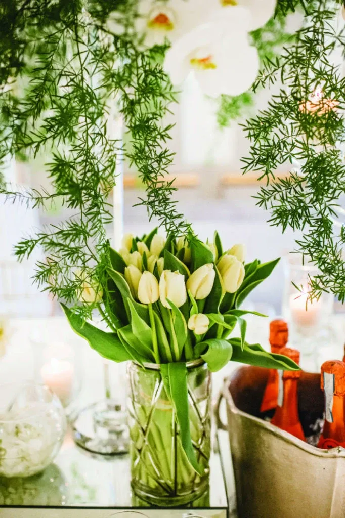 The image showcases an elegant floral arrangement by Alain Simon Fleurs, a wedding florist featured on Mariage Elegant, Canada. A vase filled with cream-colored tulips is prominently displayed, surrounded by lush green leaves. Hanging greenery cascades from above, adding a natural, romantic touch typical of wedding decor. In the background, softly blurred, are candles and additional glass containers, suggesting a sophisticated table setting perfect for wedding celebrations.