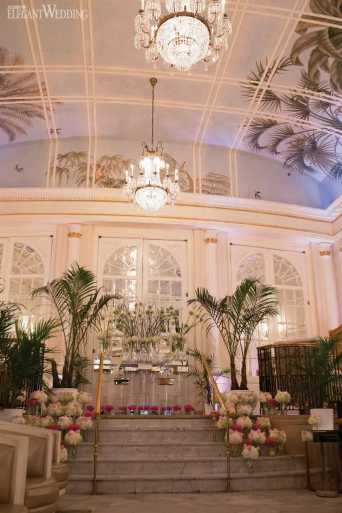 The image showcases a luxurious wedding venue beautifully decorated by Alain Simon Fleurs, a renowned wedding florist, under the vendor category of wedding florists. The grand staircase is adorned with elegant floral arrangements featuring white and pink blooms in tall glass vases, accompanied by lush greenery. Crystal chandeliers hang from the ornate ceiling, casting a warm glow over the space. The background features delicate wall frescoes and arched doorways, adding to the sophisticated ambiance perfect for an elegant wedding celebration.