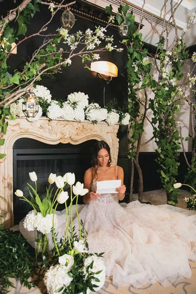 In the image, a bride in an elegant white wedding gown is seated by an ornate, carved fireplace adorned with lush greenery and white floral arrangements. This setting, styled by Alain Simon Fleurs, a renowned wedding florist in Canada, features tall, graceful branches with blooming white flowers arching over the scene, creating a romantic and serene atmosphere. White tulips and other floral decorations are artistically placed around, enhancing the wedding-themed décor in a sophisticated manner. The bride holds a letter, adding a personal and intimate touch to the wedding scene. This beautifully arranged setup showcases the exquisite floral artistry typical of Alain Simon Fleurs, making it an ideal choice for couples seeking elegant floral designs for their special day.