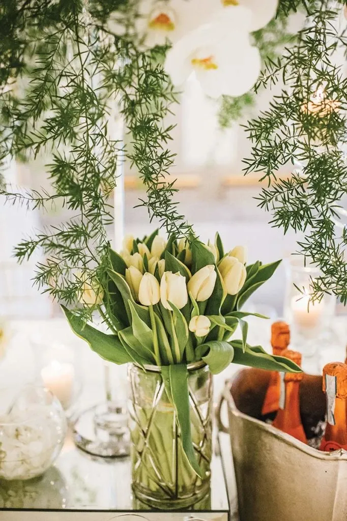 The image features a beautiful floral arrangement by Alain Simon Fleurs, a renowned wedding florist. The arrangement consists of elegant white tulips displayed in a patterned glass vase, surrounded by cascading greenery. The setting conveys a sophisticated and romantic ambiance, ideal for wedding decor. In the background, there are soft glowing candles and a metal bucket with bottles, suggesting a celebratory event. This image highlights the exquisite craftsmanship and attention to detail offered by Alain Simon Fleurs for weddings.