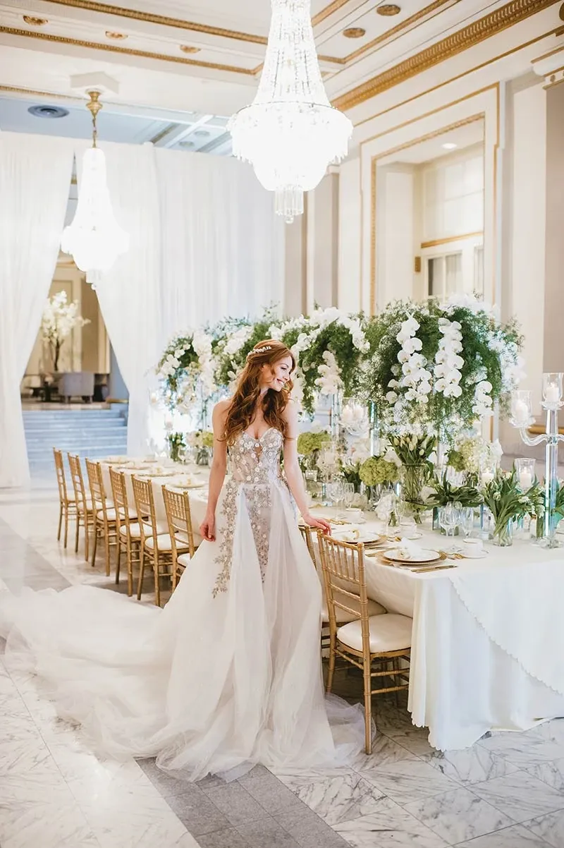 A bride stands elegantly beside a beautifully arranged wedding reception table, showcasing the floral artistry of Alain Simon Fleurs, a renowned wedding florist in Canada. The table is adorned with lush, cascading white orchids and greenery, creating a stunning centerpiece. Tall glass candle holders with lit candles add a touch of romance, while the opulent chandeliers overhead reflect the luxurious setting. The room is decorated with a classic and sophisticated aesthetic, featuring gold chiavari chairs and a marble floor, perfect for an elegant wedding celebration.