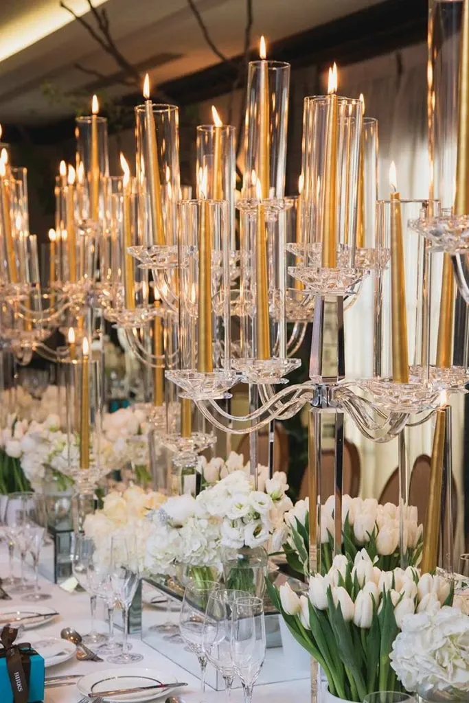 The image showcases an elegant wedding floral arrangement by Alain Simon Fleurs, a wedding florist listed on Mariage Elegant in Canada. Tall, clear candle holders with lit tapered candles illuminate a luxurious table setting. The table features abundant white flowers, including tulips and roses, beautifully arranged in sleek glass vases. The sophisticated design creates a romantic ambiance suitable for an upscale wedding reception.