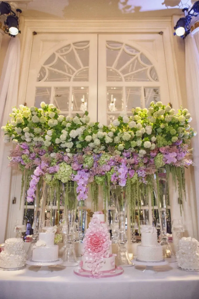 In this image from Mariage Elegant, showcasing Alain Simon Fleurs, a wedding florist in Canada, a stunning floral arrangement is on display. The backdrop features an elegant arch of white and pale green hydrangeas, intermingled with cascading purple orchids, creating a lush and romantic aesthetic. Below the florals, an array of elegant wedding cakes, including a centerpiece cake adorned with a large pink flower, is arranged on a table. The setting exudes a sense of luxury and sophistication, perfect for an upscale wedding celebration.
