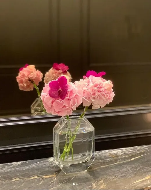 This image features a floral arrangement by Alain Simon Fleurs, a distinguished wedding florist in Canada, featured on the Mariage Elegant website. The arrangement consists of delicate pink and magenta flowers, elegantly displayed in a clear, geometric vase. The flowers are neatly organized, showcasing the florist’s attention to detail and artistry in creating romantic wedding decorations. The arrangement is set against a dark, reflective background, highlighting the vibrant colors and adding a sophisticated touch to the overall presentation.