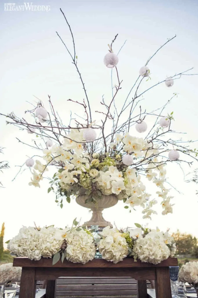 The image showcases a beautiful floral arrangement by Alain Simon Fleurs, categorized under wedding florists, displayed on Mariage Elegant's vendor listing. The centerpiece features a stunning array of white blossoms, including lush hydrangeas and elegant orchids, artistically arranged in a large, decorative urn. Delicate branches extend upwards, decorated with small, white, spherical ornaments, adding a whimsical touch. The arrangement sits atop a wooden table adorned with a lush garland of hydrangeas, capturing the essence of sophisticated wedding décor under a clear sky.