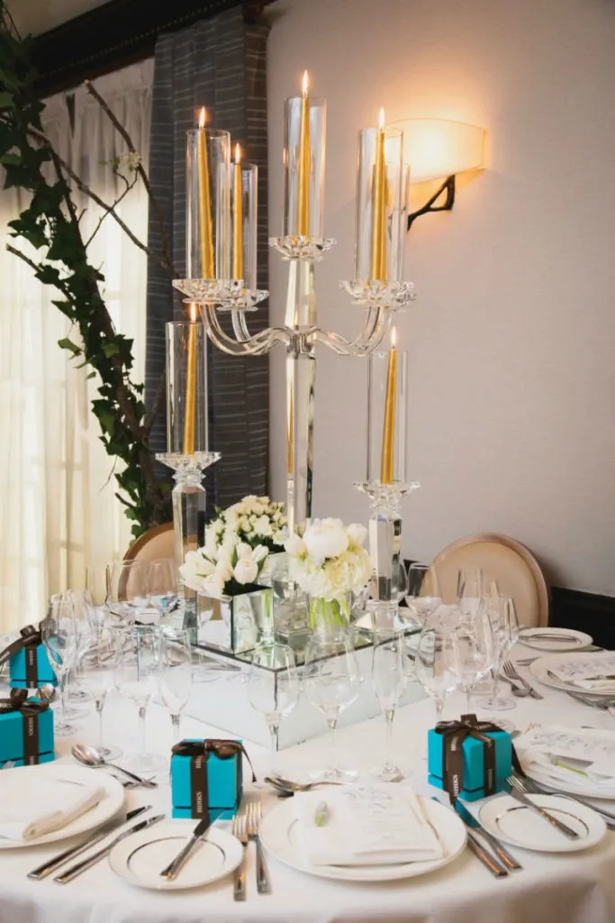 The image features an elegant wedding table arrangement by Alain Simon Fleurs, a renowned wedding florist in Canada. The centerpiece is a tall, exquisite candelabra with multiple golden candles, surrounded by a lush arrangement of white roses and green foliage. The table is set with fine china, crystal glasses, and neatly arranged silverware. Small, elegantly wrapped turquoise gift boxes are placed at each setting, adding a touch of sophistication. The backdrop features soft, ambient lighting and a glimpse of sheer drapes, creating a romantic atmosphere perfect for a wedding celebration.