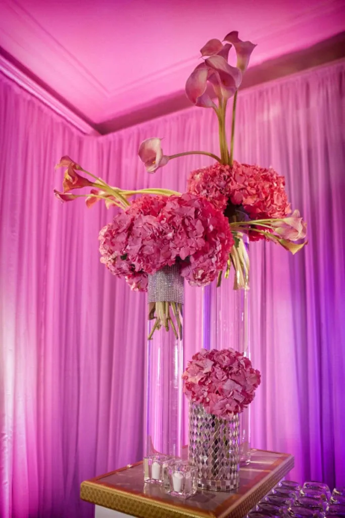 The image showcases a stunning floral arrangement by Alain Simon Fleurs, a vendor in the Wedding Florists category for Mariage Elegant, Canada. The display features tall, sleek glass vases filled with vibrant pink hydrangeas and calla lilies, elegantly arranged for a wedding setting. The background is adorned with soft, flowing pink drapes, creating a romantic and sophisticated atmosphere. The vases are embellished with sparkling rhinestones, adding a touch of glamour and elegance to the floral arrangement. Perfect for an exquisite wedding celebration, the design exemplifies high-end floral artistry.