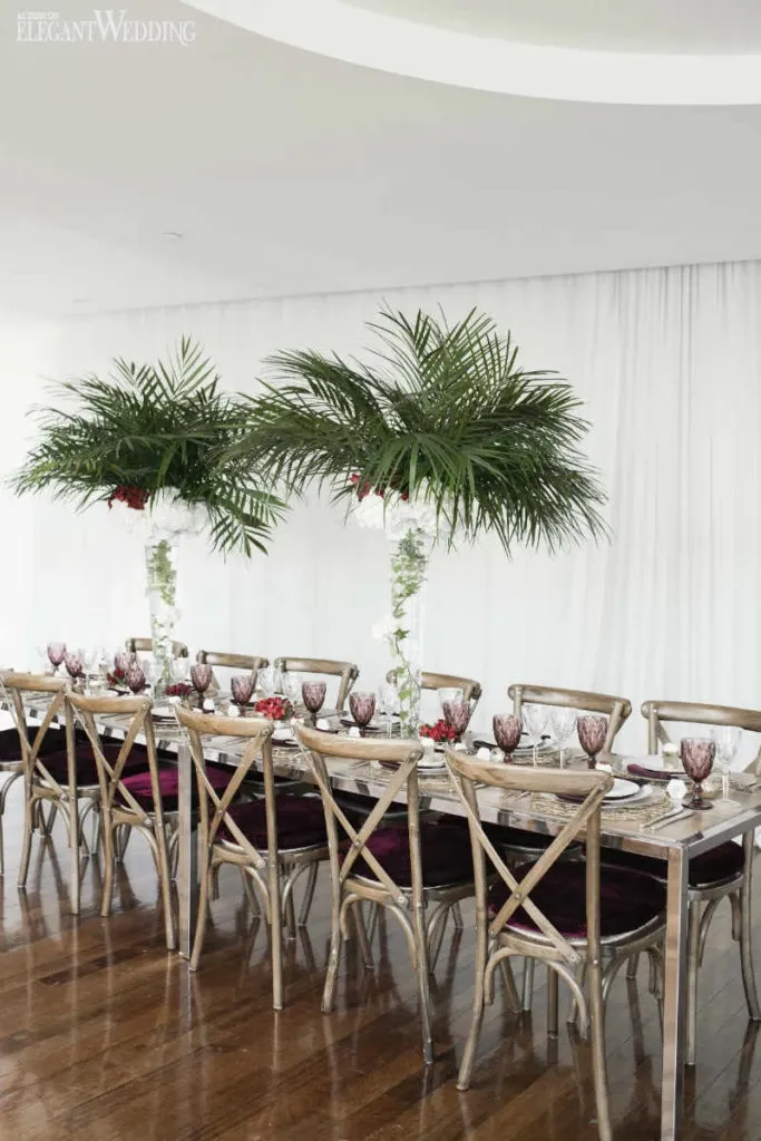 The image showcases an exquisite wedding reception table arrangement by Alain Simon Fleurs, a wedding florist in Canada. The long wooden table is elegantly set with high-back wooden chairs and decorated with tall floral centerpieces featuring lush green palm leaves, creating a tropical and sophisticated ambiance. The table is adorned with deep red flowers, clear glassware, and intricately patterned white place settings, set against a backdrop of sheer white curtains that add a touch of elegance to the venue. This setup perfectly highlights the artistry and skill of Alain Simon Fleurs in creating stunning wedding floral designs.