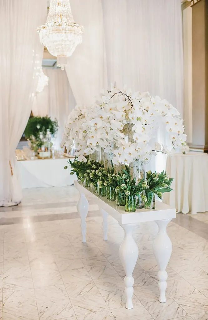 The image showcases an elegant floral arrangement by Alain Simon Fleurs, a wedding florist featured on Mariage Elegant, Canada. The display consists of a series of lush white orchids cascading from tall vases, set upon a chic white table with turned legs. Surrounding the orchids are smaller vases filled with green foliage, adding a rich contrast to the white blooms. The setup is placed in a sophisticated venue, with a luxurious chandelier hanging above and flowing white drapes adorning the background, embodying a classy wedding ambiance.