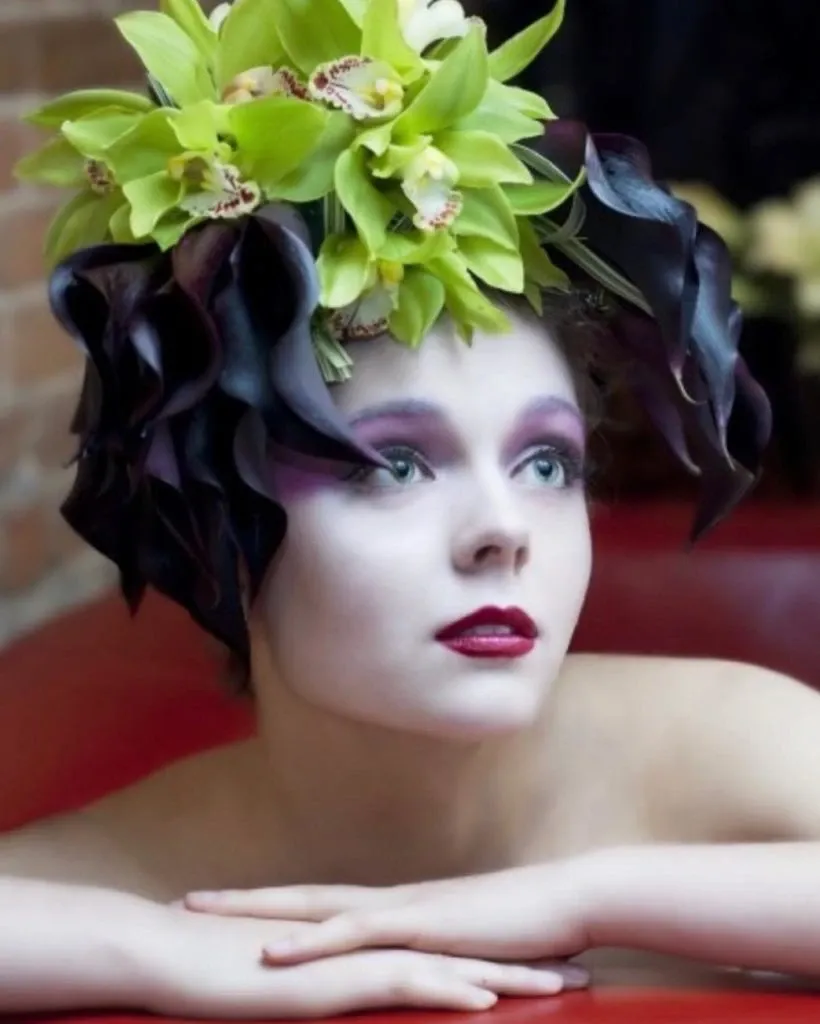 In the image, a model is showcasing a striking floral headpiece by Alain Simon Fleurs, a vendor specializing in wedding florists at Mariage Elegant, Canada. The headpiece features vibrant green orchids elegantly paired with dark, dramatic calla lilies, creating a stunning and avant-garde look. The model's makeup complements the arrangement with soft purple eyeshadow and bold red lipstick, adding to the overall artistic and sophisticated aesthetic ideal for a unique wedding style.