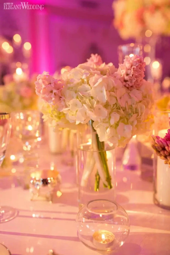 The image showcases a stunning floral arrangement by Alain Simon Fleurs, a premier wedding florist in Canada. The centerpiece features lush white and pink hydrangeas, beautifully arranged in a clear glass vase, set on an elegantly decorated wedding table. The table is adorned with small, glowing candles and reflective glassware, creating a romantic ambiance. Soft, purple lighting adds a touch of sophistication to the setting, embodying the elegant wedding theme promoted by Mariage Elegant.