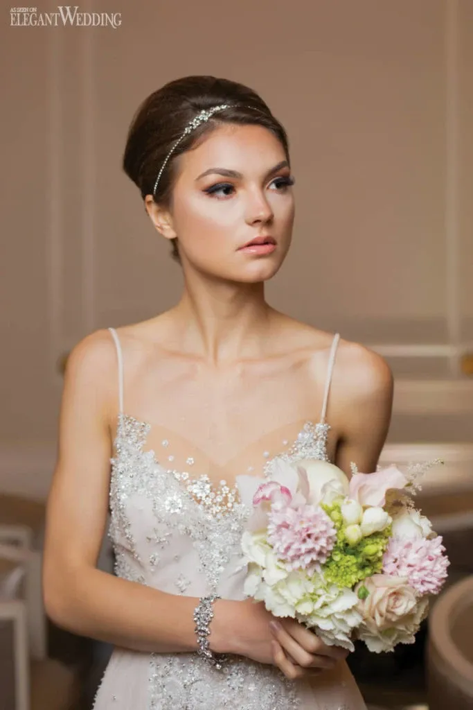 A bride holding a bouquet made by Alain Simon Fleurs, a wedding florist, is showcased in this image. The bouquet features a mix of soft pink and white flowers, including roses and hydrangeas, accented with green foliage. The bride is wearing a delicate, embellished gown with fine detailing, and a simple crystal headband adorns her hair. The setting appears elegant and sophisticated, aligning with the style of Mariage Elegant in Canada.