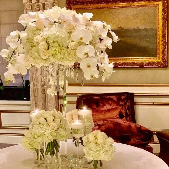 The image showcases an elegant floral arrangement by Alain Simon Fleurs, categorized under Wedding Florists, for Mariage Elegant in Canada. A tall centerpiece features cascading white orchids and roses atop a clear glass vase adorned with hanging crystal strands. Surrounding the centerpiece are smaller arrangements of white roses and hydrangeas, complemented by the soft glow of pillar candles in glass holders. The setting suggests a refined and luxurious wedding ambiance, enhanced by an opulent room with rich furnishings and an ornate painting in the background.