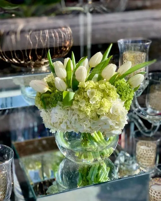 The image features an elegant floral arrangement by Alain Simon Fleurs, a wedding florist listed on Mariage Elegant, Canada. This arrangement showcases a delicate blend of white tulips, lush green hydrangeas, and vibrant greenery, all artistically displayed in a clear glass vase. The vases are set on a reflective mirrored surface, enhancing the chic and sophisticated appeal. Surrounding the bouquet, there are intricately designed candle holders that add a touch of romance, making this arrangement a perfect choice for wedding decor.