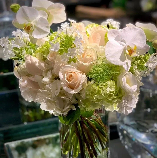 In this image, a beautifully arranged wedding bouquet by Alain Simon Fleurs, a renowned wedding florist, is featured. The bouquet includes elegant white orchids, soft peach roses, delicate hydrangeas, and lush green foliage. The flowers are artistically arranged to create a luxurious and romantic feel, perfect for a sophisticated wedding setting. The bouquet is placed against a blurred background, highlighting the expertise of the florist in creating stunning floral arrangements for special occasions. This showcases the high-quality floral design services available for weddings in Canada through Mariage Elegant.