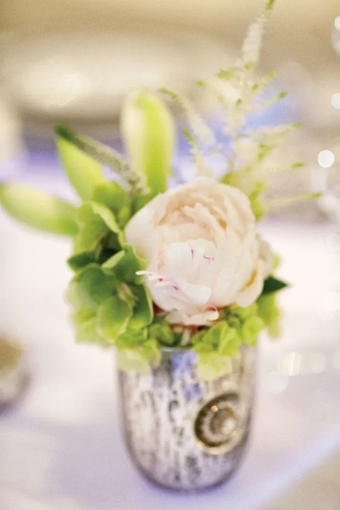 This image showcases a beautifully arranged floral centerpiece by Alain Simon Fleurs, a renowned wedding florist. The arrangement features a delicate white peony as the focal point, surrounded by vibrant green orchids and lush greenery, all elegantly placed in a textured silver vase. The soft and romantic design is perfect for enhancing the elegance of any wedding venue, reflecting the sophisticated craftsmanship of the vendor. The gentle lighting adds a warm ambiance, ideal for a wedding celebration.