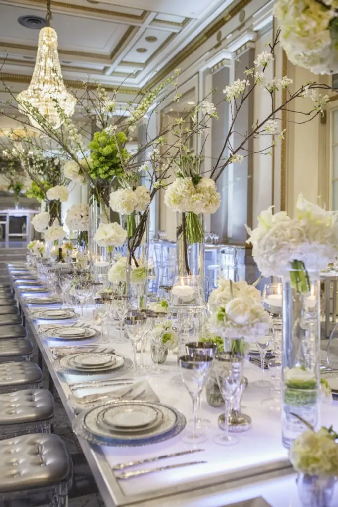 This image features a beautifully arranged wedding reception table, highlighting the artistry of Alain Simon Fleurs, a vendor in the wedding florists category. The table is elegantly adorned with tall glass vases filled with delicate white and green floral arrangements, including hydrangeas and branches, creating a sophisticated and natural ambiance. Each setting is meticulously arranged with crystal glassware, silver chargers, and fine china. Overhead, a large, luxurious chandelier adds a touch of glamour to the setting. The image perfectly captures the elegant atmosphere suitable for an upscale wedding event in Canada.