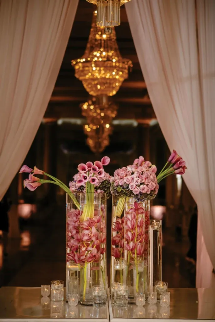 The image showcases a luxurious floral arrangement by Alain Simon Fleurs, a wedding florist listed on Mariage Elegant. The arrangement features tall, clear glass vases filled with elegant pink calla lilies and roses, beautifully displayed beneath an opulent chandelier. The backdrop includes soft, flowing white drapes, contributing to the sophisticated and romantic ambiance perfect for a wedding setting.