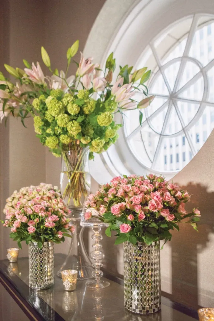 The image features a beautifully arranged display by Alain Simon Fleurs, a prominent wedding florist in Canada. Three elegant vases are positioned on a glass table beneath a circular window, each filled with lush floral arrangements. The central bouquet in a tall, clear vase is composed of green hydrangeas and pink lilies, adding height and grandeur. Flanking it are two smaller vases with intricate geometric patterns, each filled with pink roses and white baby's breath, offering a romantic touch. Soft candlelight from two gold, votive candle holders enhances the elegant atmosphere, making it an ideal setting for a wedding reception or ceremony.