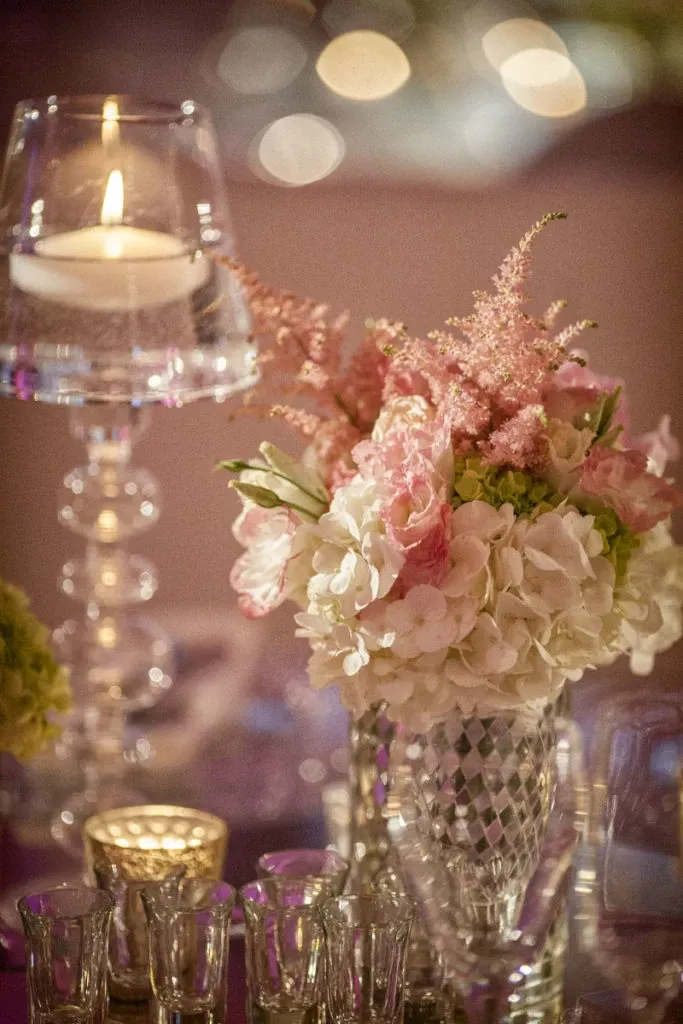The image showcases a beautifully arranged floral centerpiece by Alain Simon Fleurs, categorized under Wedding Florists, displayed for Mariage Elegant in Canada. The arrangement features a delicate mix of white hydrangeas and soft pink flowers, elegantly presented in an intricately cut crystal vase. A tall, elegant glass candlestick with a lit candle adds a romantic glow to the scene, highlighting the exquisite craftsmanship of the floral design. The setting exudes a luxurious and elegant ambiance, perfect for a sophisticated wedding atmosphere.