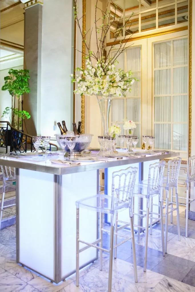 The elegant floral arrangement by Alain Simon Fleurs, a premier wedding florist, is prominently displayed on a high-lit table at a sophisticated wedding venue in Canada. A tall, clear vase holds a lush bouquet of white flowers and delicate branches, adding a touch of natural beauty to the setting. Surrounding the arrangement, transparent chairs and an elegantly set table with champagne glasses and an ice bucket create a luxurious atmosphere, perfect for an upscale wedding celebration featured on Mariage Elegant.