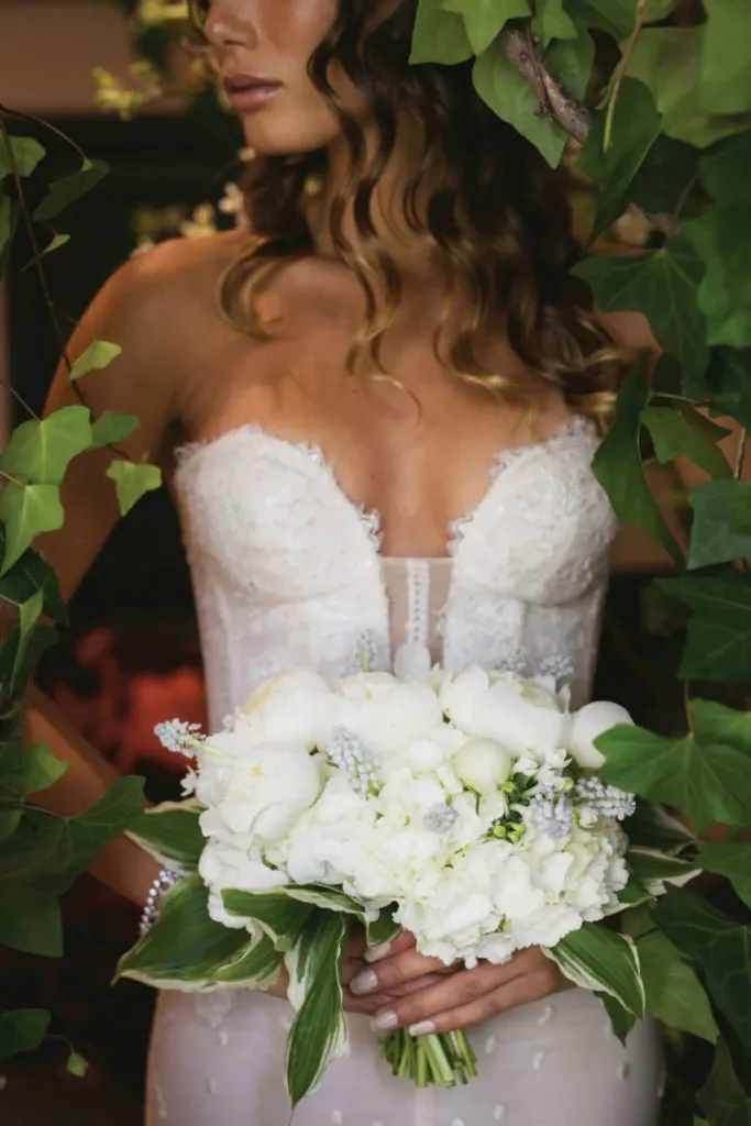 A bride, surrounded by lush greenery, holds a stunning bouquet of white flowers, showcasing the exceptional floral artistry of Alain Simon Fleurs, a leading vendor in the Wedding Florists category. Her elegant lace wedding dress complements the delicate arrangement, which includes roses and other fresh blossoms, ideal for sophisticated wedding ceremonies in Canada.