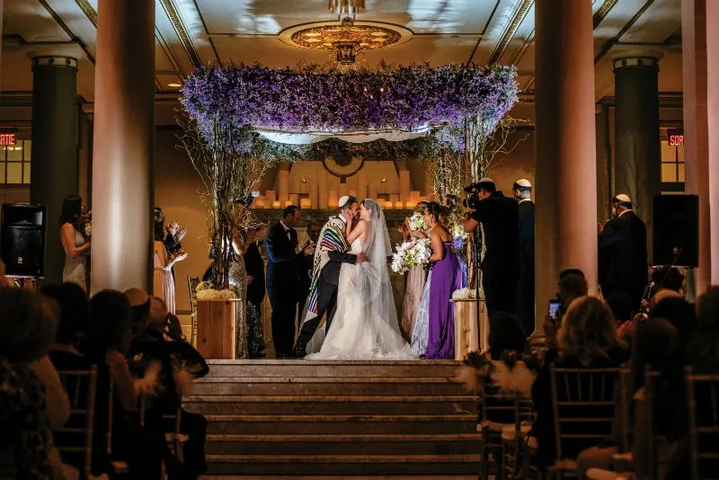 The image features a beautifully decorated wedding venue with a floral arch created by Alain Simon Fleurs, a renowned vendor in the Wedding Florists category. The arch, adorned with purple and white flowers, serves as a stunning backdrop for the bride and groom sharing a kiss during their ceremony. The setting is elegant, with tall columns and an ornate ceiling, creating a romantic atmosphere. Guests are seated on either side, watching the couple in a moment of joy. The craftsmanship of the floral arrangement adds an exquisite touch to the wedding celebration.
