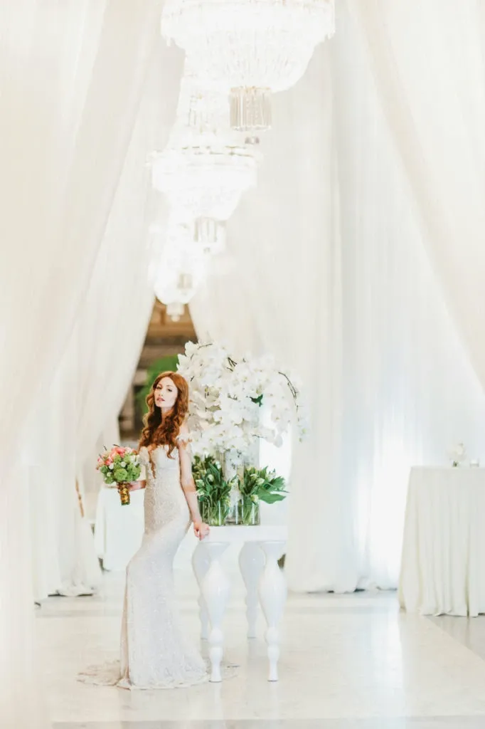 A bride stands elegantly in a beautifully decorated venue, holding a vibrant bouquet of flowers created by Alain Simon Fleurs, a distinguished vendor in the Wedding Florists category. The setting features luxurious white drapes and grand chandeliers, providing a sophisticated backdrop. Next to the bride, large arrangements of lush white orchids and greenery are displayed on a chic white pedestal table, showcasing the florist’s expertise in creating stunning wedding arrangements. The overall ambiance exudes elegance and style, making it perfect for a high-end wedding celebration.